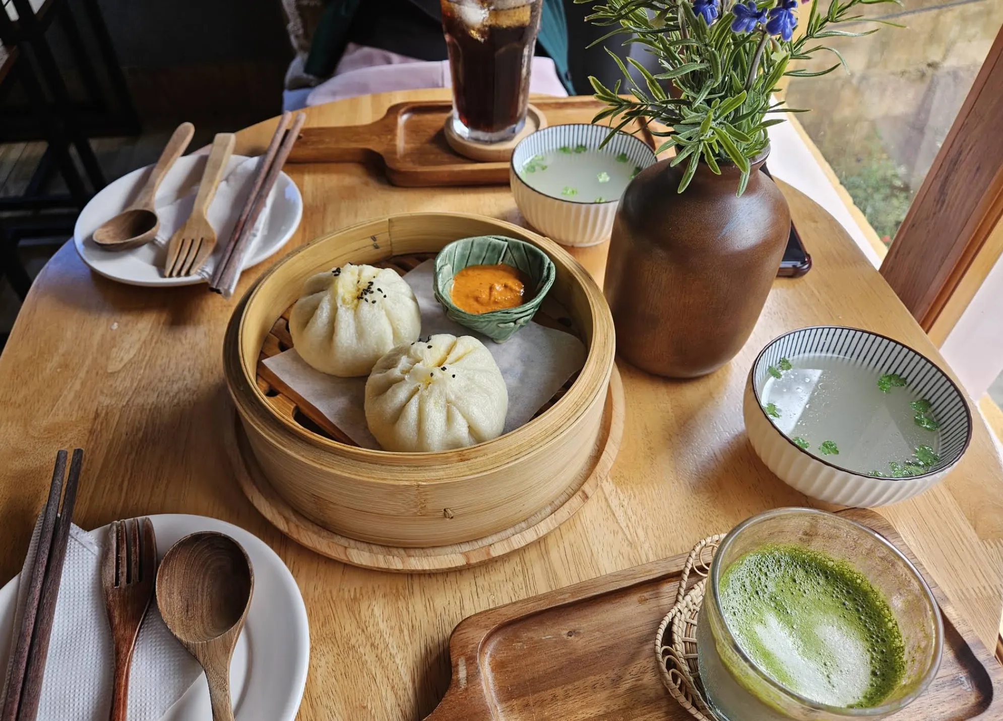 Two steamed bao in a bamboo steamer with dipping sauce, clear soup and a matcha latte
